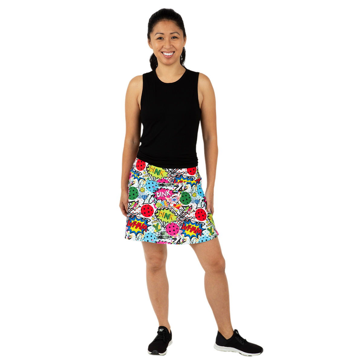 A smiling woman with a ponytail stands on a white background, wearing Queen of the Court's Dink Dink Wham Swing Skirt—a colorful pickleball skirt with comic book-style graphics—paired with a sleeveless black top and black sneakers.