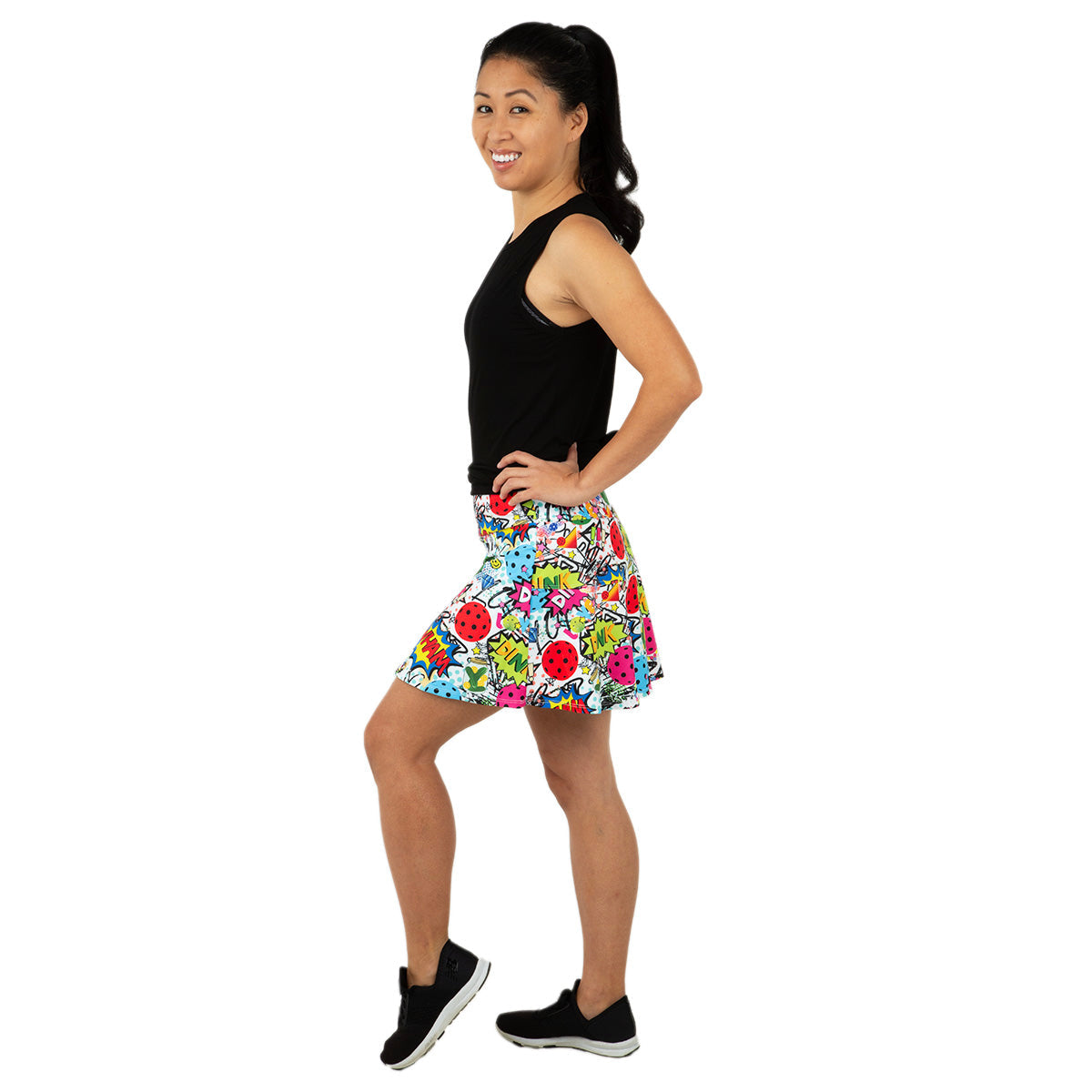 A woman with dark hair in a ponytail smiles and stands sideways, wearing the Queen of the Court Dink Dink Wham Swing Skirt with bold comic-inspired graphics, a sleeveless black top, and black sneakers with white soles against a white background.