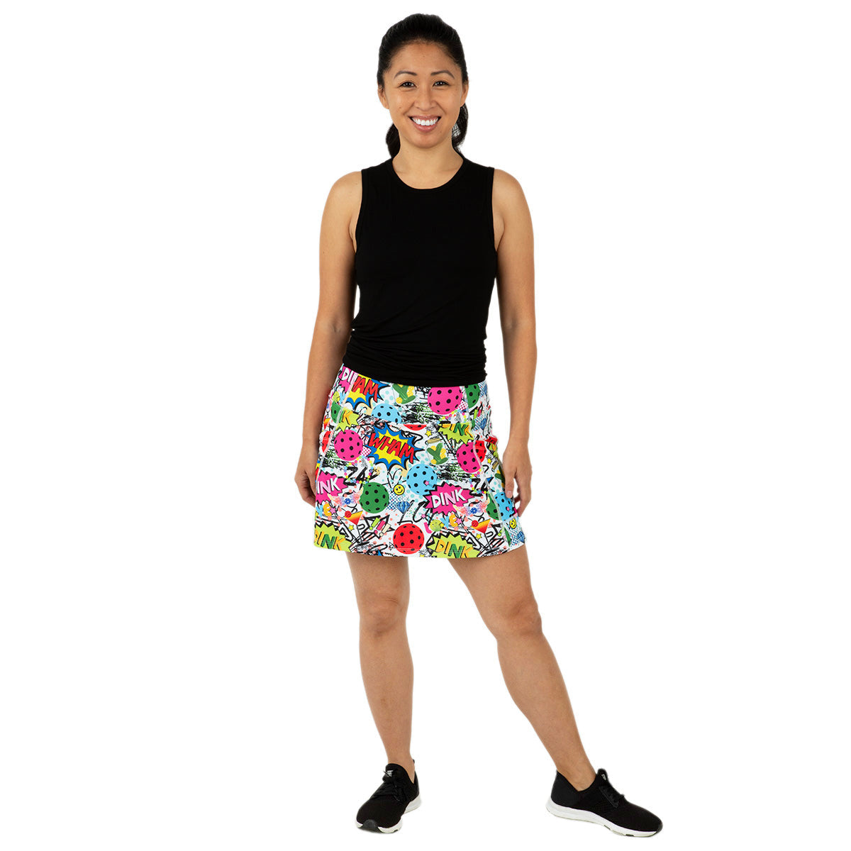 A woman smiles against a white background, wearing the Queen of the Court Dink Dink Wham Endurance 15" Skirt with comic book-style graphics and side pockets, a sleeveless black top, and black sneakers with white soles. One leg is slightly bent.