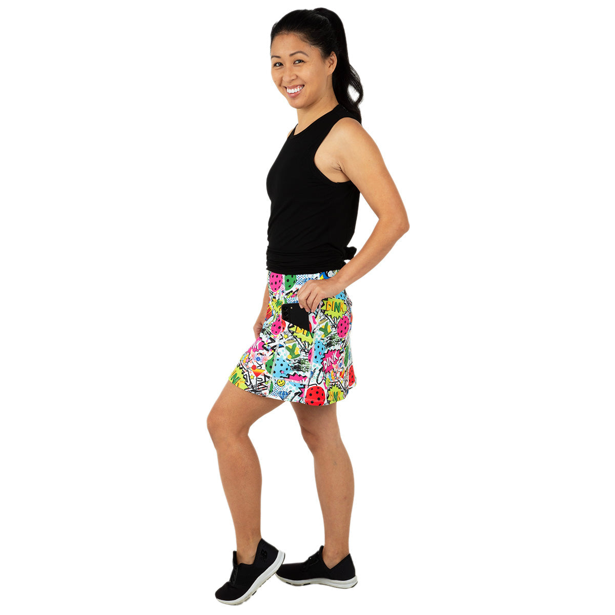 A woman stands smiling in a side pose against a white background, wearing the Queen of the Court Dink Dink Wham Endurance 15" Skirt with side pockets, a sleeveless black top, black slip-on sneakers, and her dark hair in a ponytail.