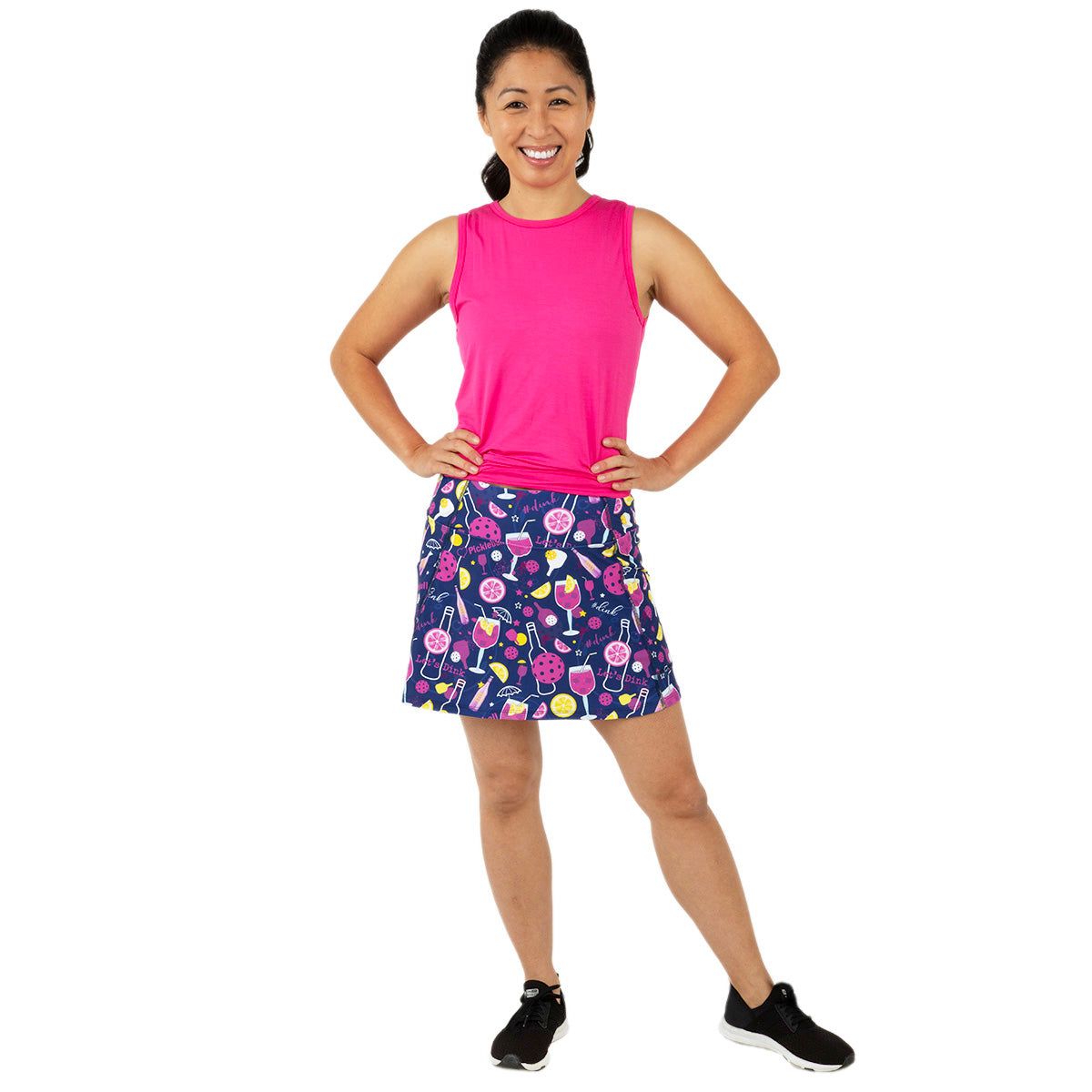 A smiling woman stands against a white background, hands on hips, wearing Queen of the Court's Dinks and Drinks Endurance 15" Skirt—navy with colorful tennis-themed patterns—a bright pink sleeveless top, black athletic shoes, and her hair in a ponytail.