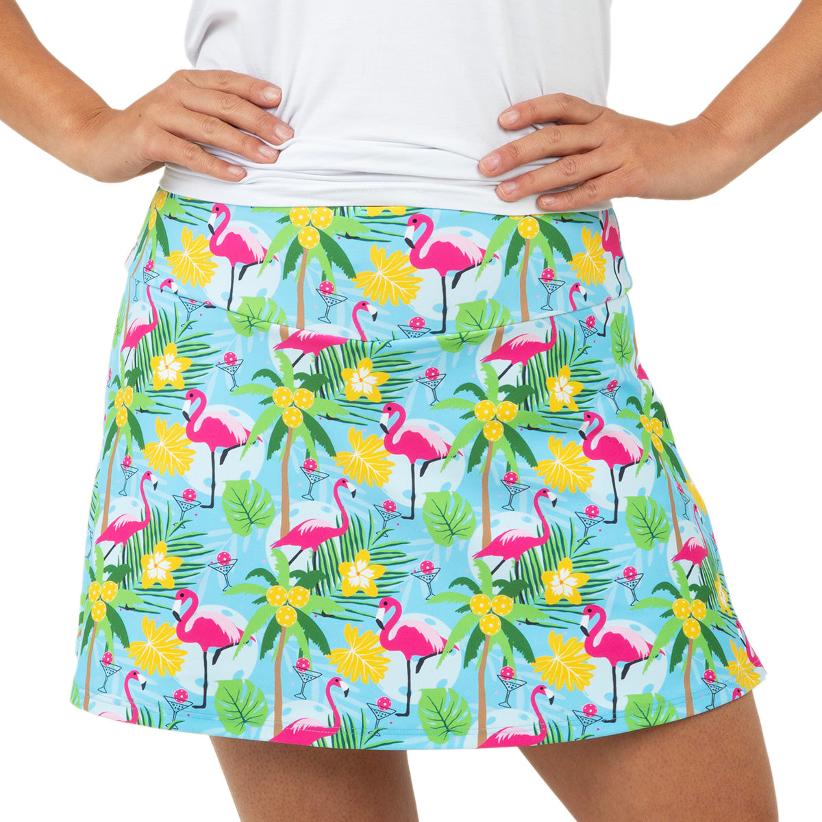 Only the torso and skirt are visible as a person stands hands on hips, wearing a white shirt and the Queen of the Court Blue Pickleball and Flamingos Swing Skirt with a tropical pattern of pink flamingos, yellow hibiscus, and green palm leaves on light blue.