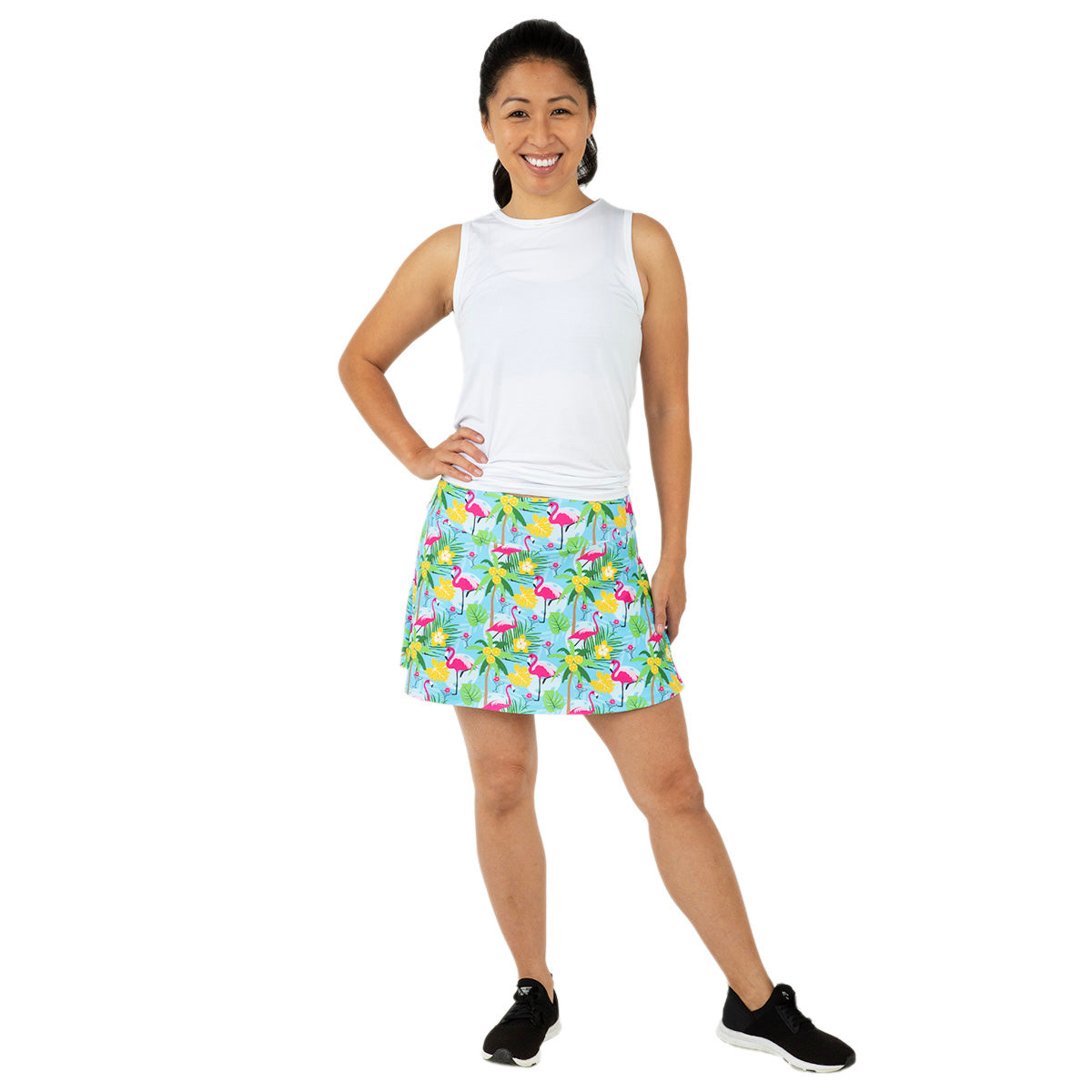 A smiling woman stands against a white background, wearing the Queen of the Court Blue Pickleball and Flamingos Swing Skirt, a sleeveless white top, and black athletic shoes. Her hair is in a ponytail as she faces the camera with her left hand on her hip.