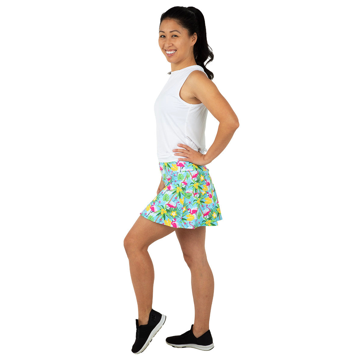 A smiling woman stands in profile against a white background, wearing a sleeveless white top, black slip-on sneakers, and the Queen of the Court Blue Pickleball and Flamingos Swing Skirt. Her dark hair is tied back in a high ponytail as she poses with one leg lifted.