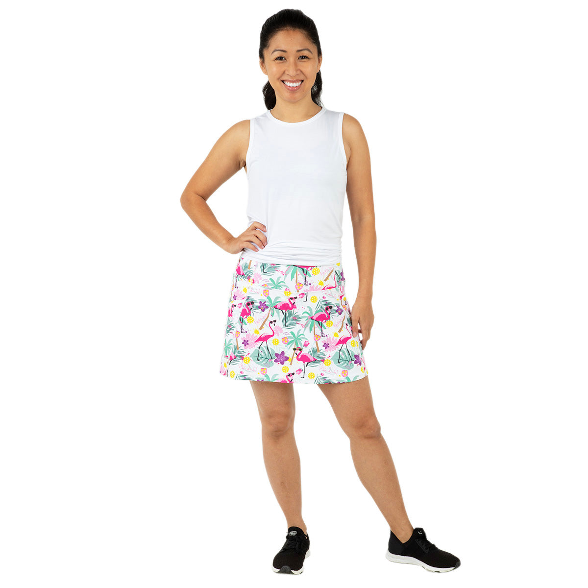 A woman smiles with one hand on her hip, wearing a sleeveless white top and the Queen of the Court White Pickleball and Flamingos Endurance 15" Skirt with black sneakers. Her dark hair is in a ponytail as she poses against a plain white background.
