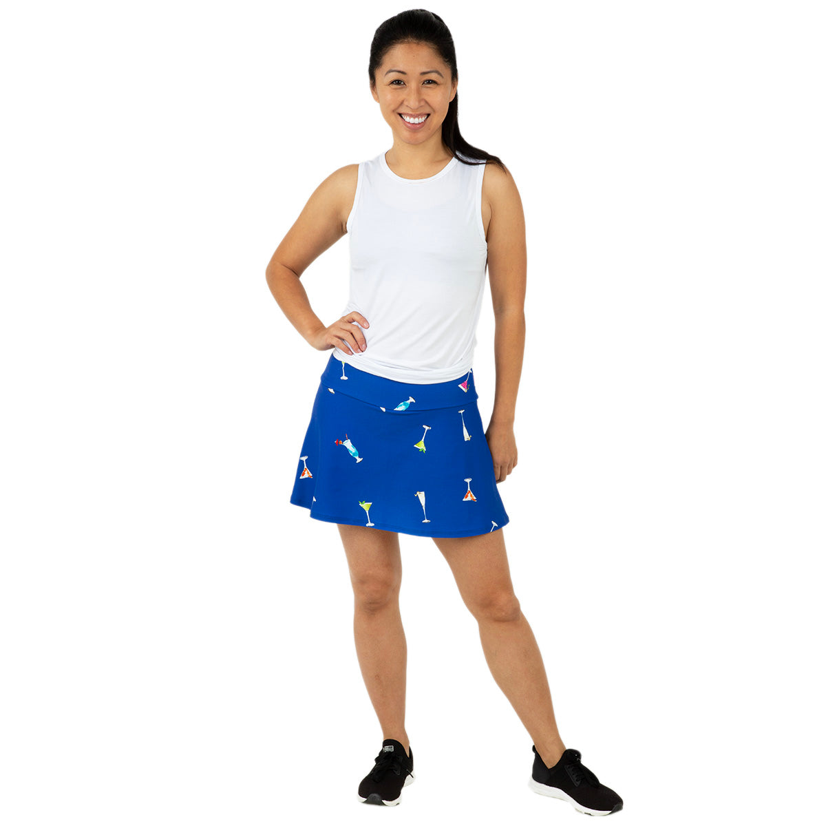 A smiling woman stands against a white background, wearing a white sleeveless top and the Queen of the Court Gin Fizz Swing Skirt, featuring colorful cocktail glass prints. Her look is completed with black athletic shoes and her long dark hair tied back.