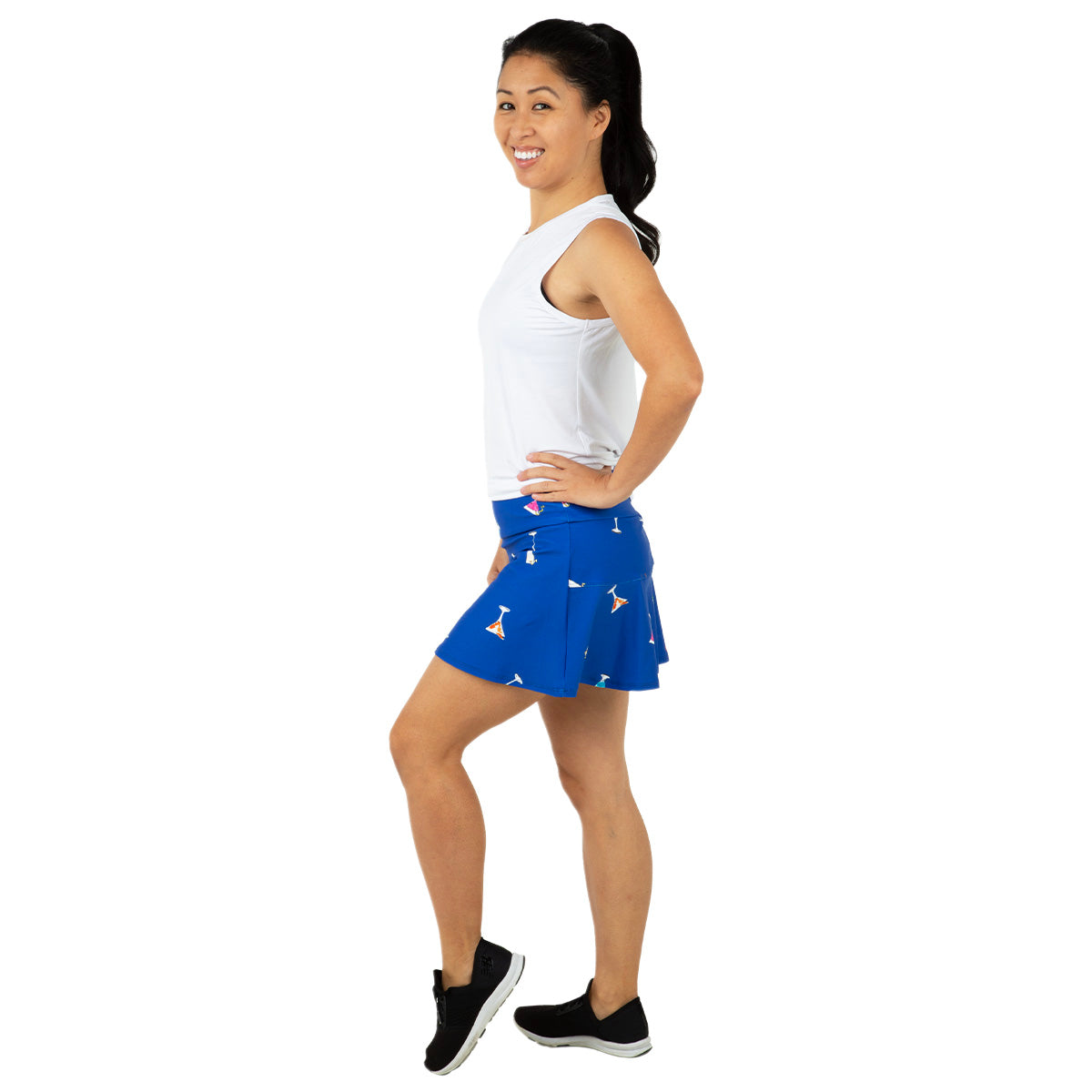 A woman stands sideways, smiling with one hand on her hip, wearing a sleeveless white top, black sneakers, and the Queen of the Court Gin Fizz Swing Skirt—a blue tennis skirt with a subtle pattern. Her dark hair is in a ponytail against a plain white background.
