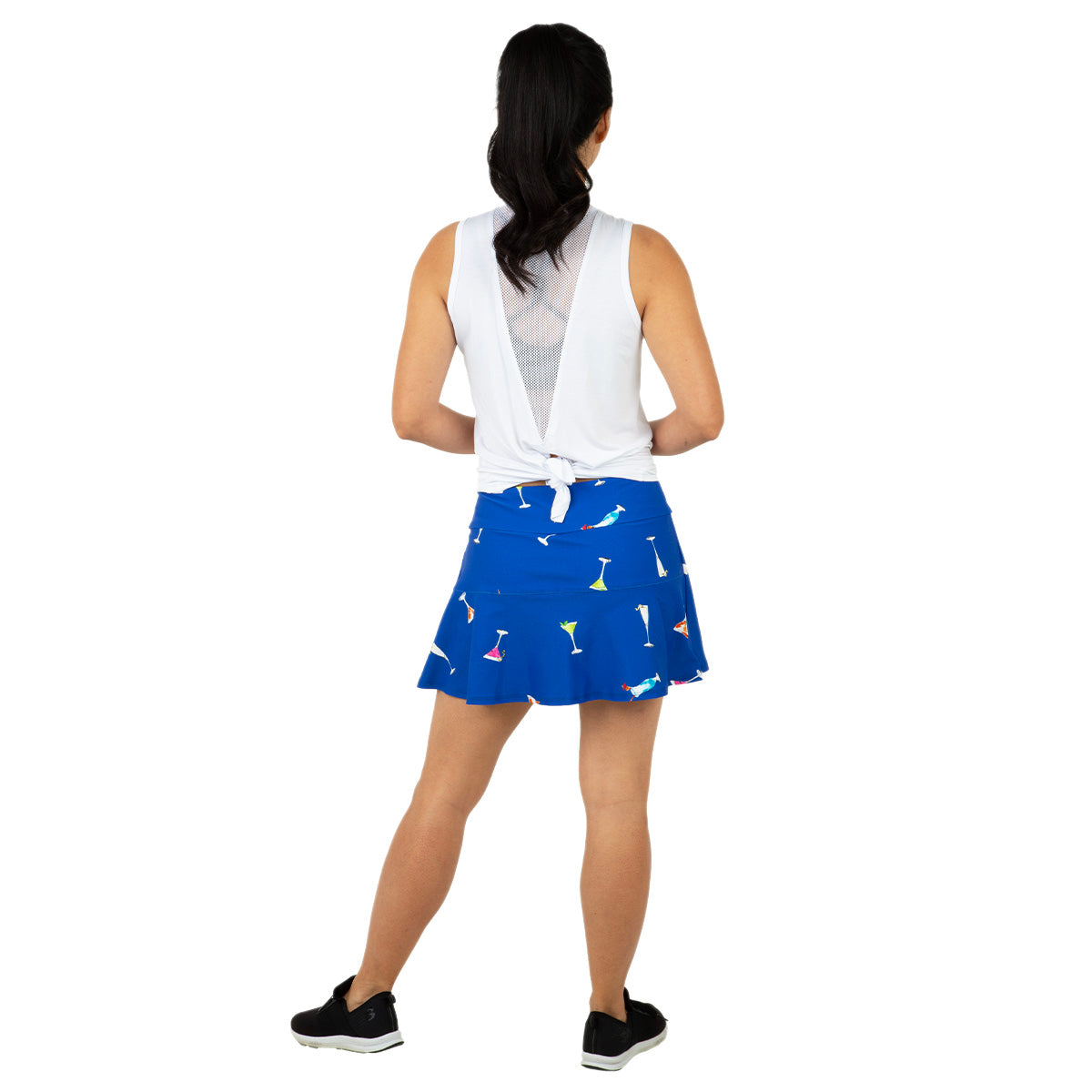 A woman with black hair in a ponytail faces away, wearing Queen of the Court's Gin Fizz Swing Skirt—a blue tennis skirt with colorful cocktail glass prints—and a white sleeveless top. She pairs this look with black sneakers and stands against a plain white background.