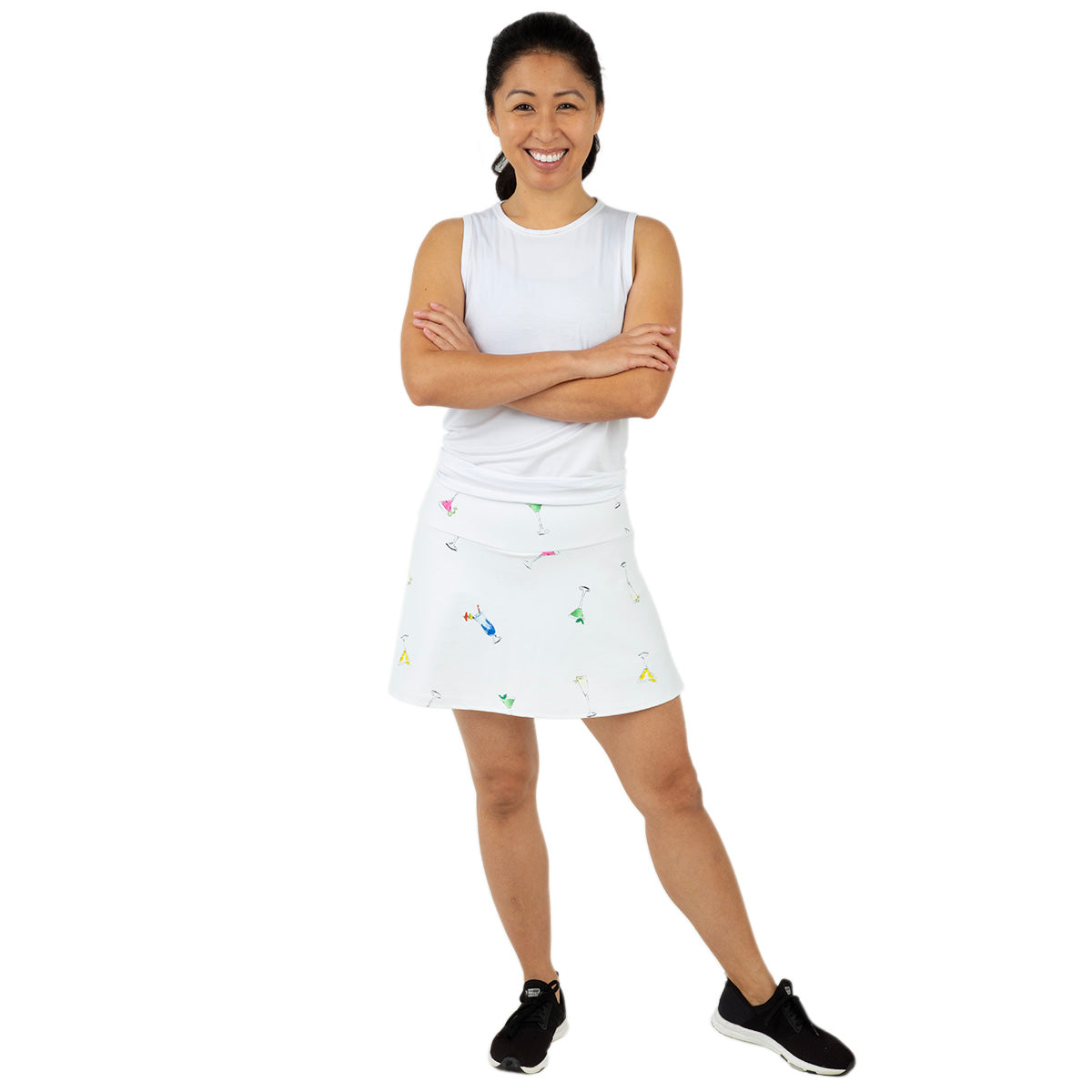 A woman with her dark hair tied back smiles confidently, wearing the Queen of the Court Gin Fizz Swing Skirt—featuring colorful prints—a sleeveless white top, and black sneakers, posed against a plain white background.