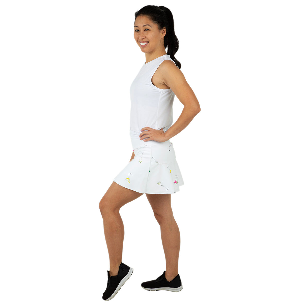 A woman with dark hair in a ponytail smiles sideways, modeling the Queen of the Court Gin Fizz Swing Skirt—a white tennis skirt with colorful patterns—paired with a sleeveless top, against a plain white background.