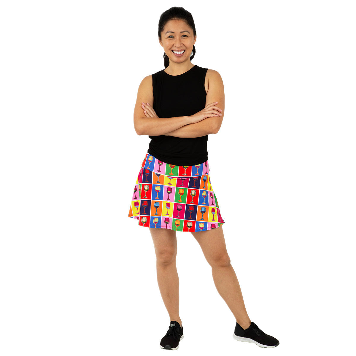 A smiling woman stands with arms crossed, wearing the Queen of the Court Red Winehol Swing Skirt—a colorful pickleball skirt with a grid of multicolored heads on white—paired with a sleeveless black top and black sneakers, against a plain white backdrop.