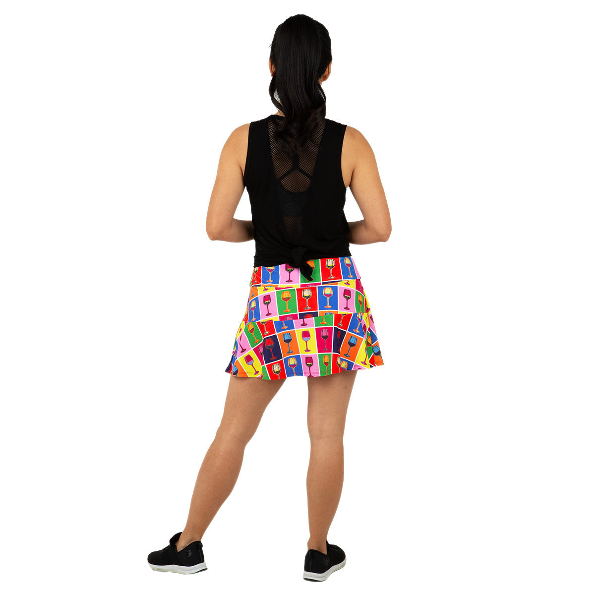 A woman faces away in a sleeveless black top with a mesh back, wearing Queen of the Court’s Red Winehol Swing Skirt, which features vibrant abstract faces. She sports black sneakers and her hair is tied back. Made in USA.
