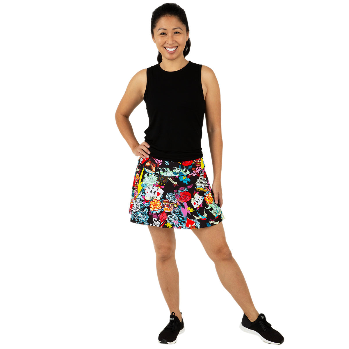 A smiling woman poses against a white background, wearing the Queen of the Court Tattoo Swing Skirt—a colorful floral A-line skirt perfect for pickleball and tennis—paired with a sleeveless black top and black athletic shoes.