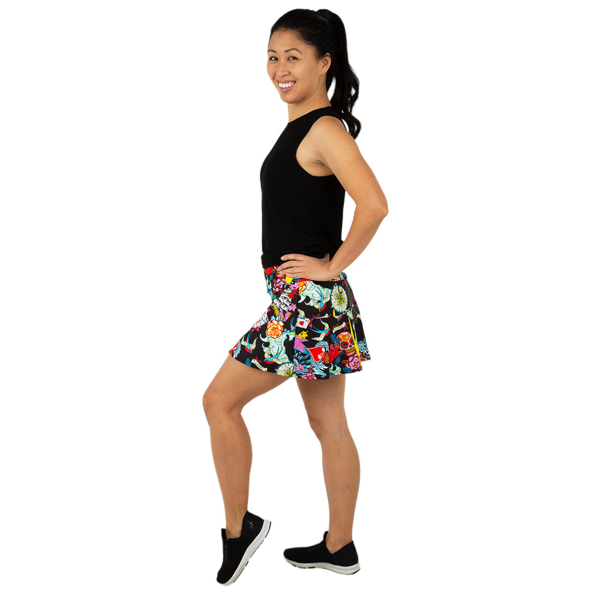 A woman stands sideways, smiling with her hands on her hips. She wears the Queen of the Court Tattoo Swing Skirt and a sleeveless black top—an ideal outfit for pickleball or tennis. Her dark hair is in a ponytail against a plain white background.