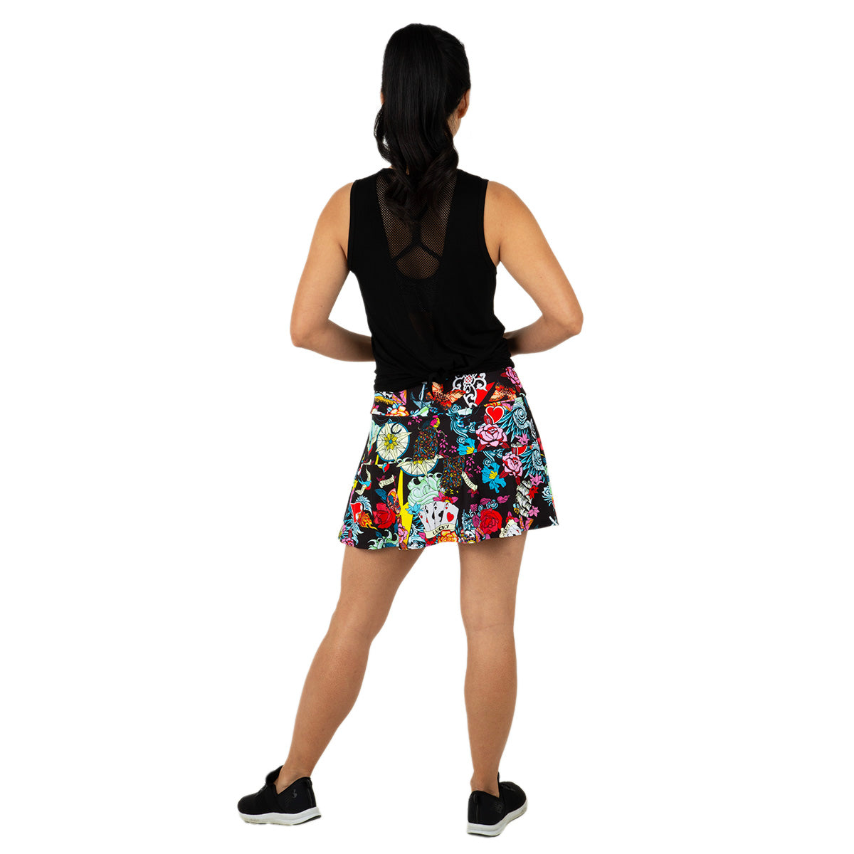 A woman stands facing away, hands on hips, wearing Queen of the Court's Tattoo Swing Skirt—a short, colorful floral skirt—and a sleeveless black top. Black sneakers and a ponytail complete her sporty look against a plain white background.