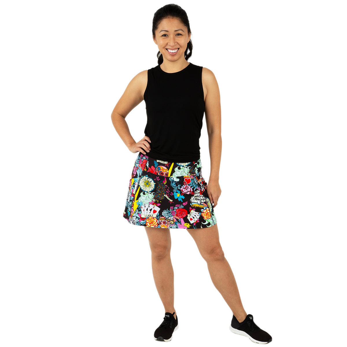 A woman with long dark hair tied back smiles against a white background, wearing a sleeveless black top, black sneakers, and the Queen of the Court Tattoo Endurance 15" Skirt featuring a colorful floral pattern. One hand is on her hip.