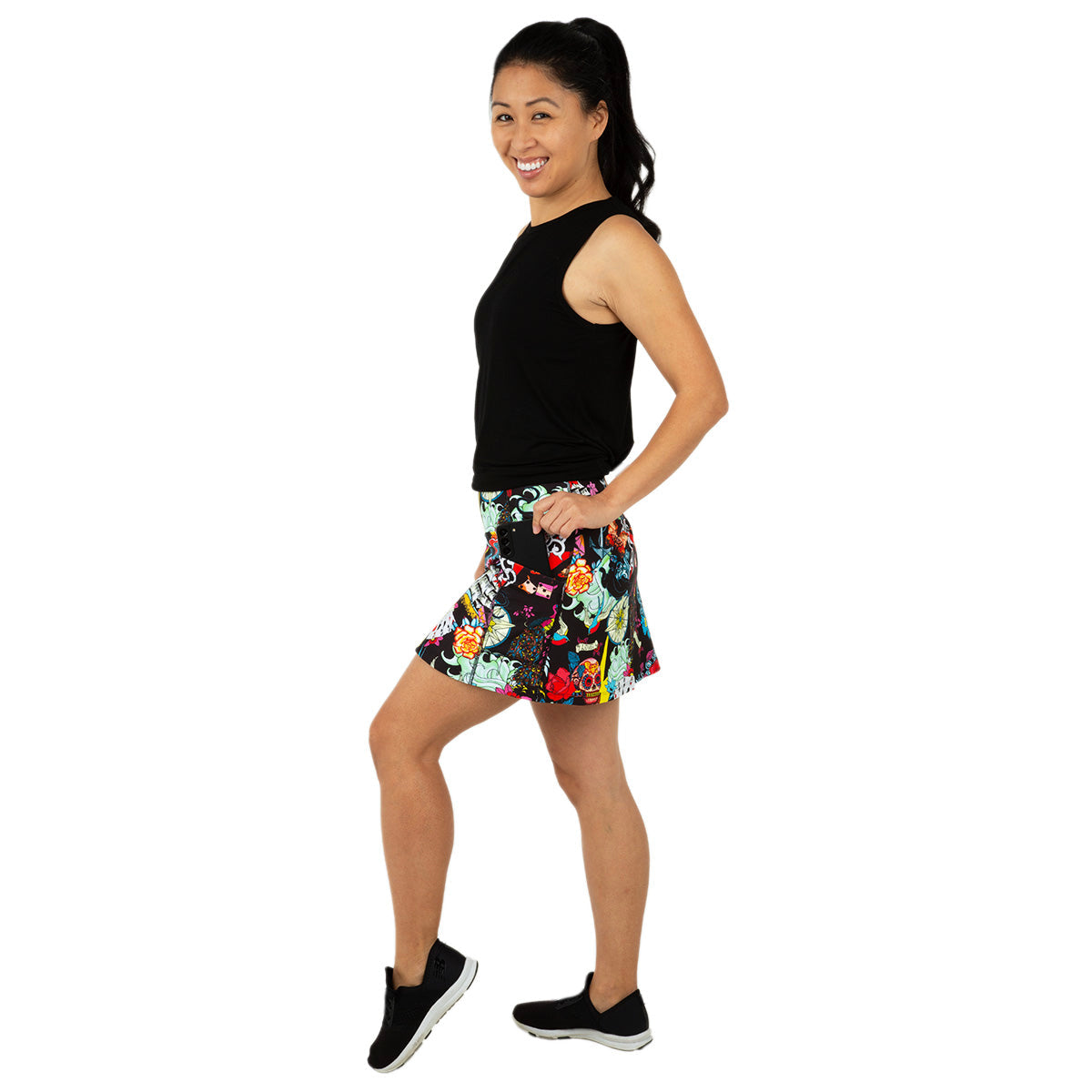 A smiling woman with a ponytail poses sideways, wearing a sleeveless black top and the Queen of the Court Tattoo Endurance 15" Skirt, a colorful floral pickleball skirt with pockets, playfully lifting one foot against a white background.