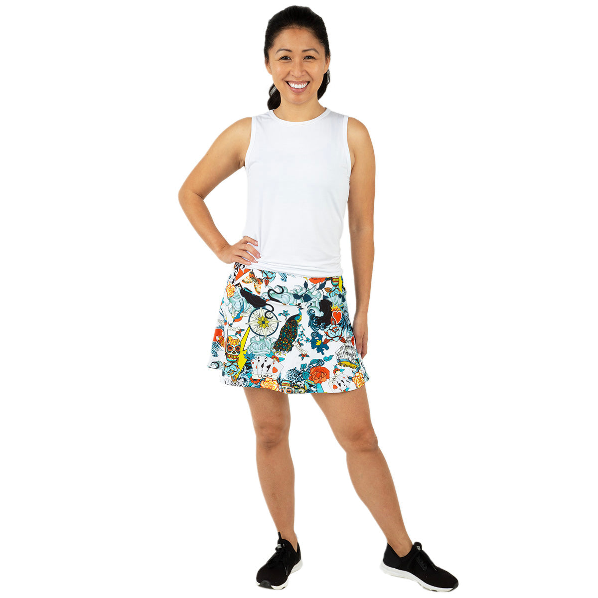 A smiling woman stands against a white background, wearing the Queen of the Court Tattoo Swing Skirt—a vibrant, pineapple-and-floral patterned performance skirt perfect for pickleball and tennis—paired with a sleeveless top and black sneakers.