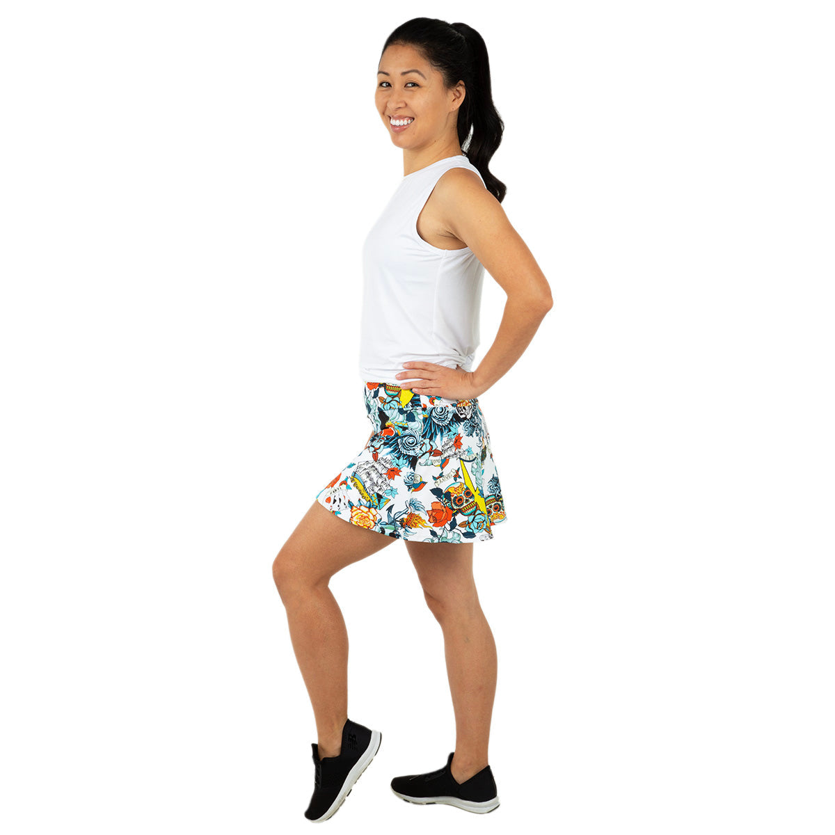 A woman stands in profile, smiling with hands on hips. She wears a sleeveless white top, Queen of the Court Tattoo Swing Skirt—ideal for pickleball and tennis—and black slip-on sneakers. Her hair is in a ponytail against a plain white background.