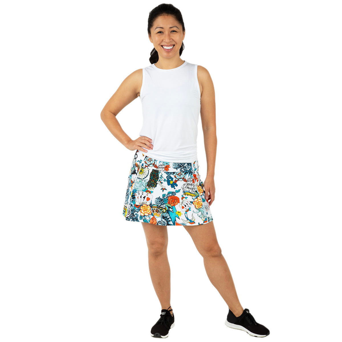 A smiling woman poses confidently against a white background, wearing the Queen of the Court Tattoo Endurance 15" Skirt—a moisture-wicking, floral and cat-patterned pickleball skirt—paired with a sleeveless white top and black athletic shoes.