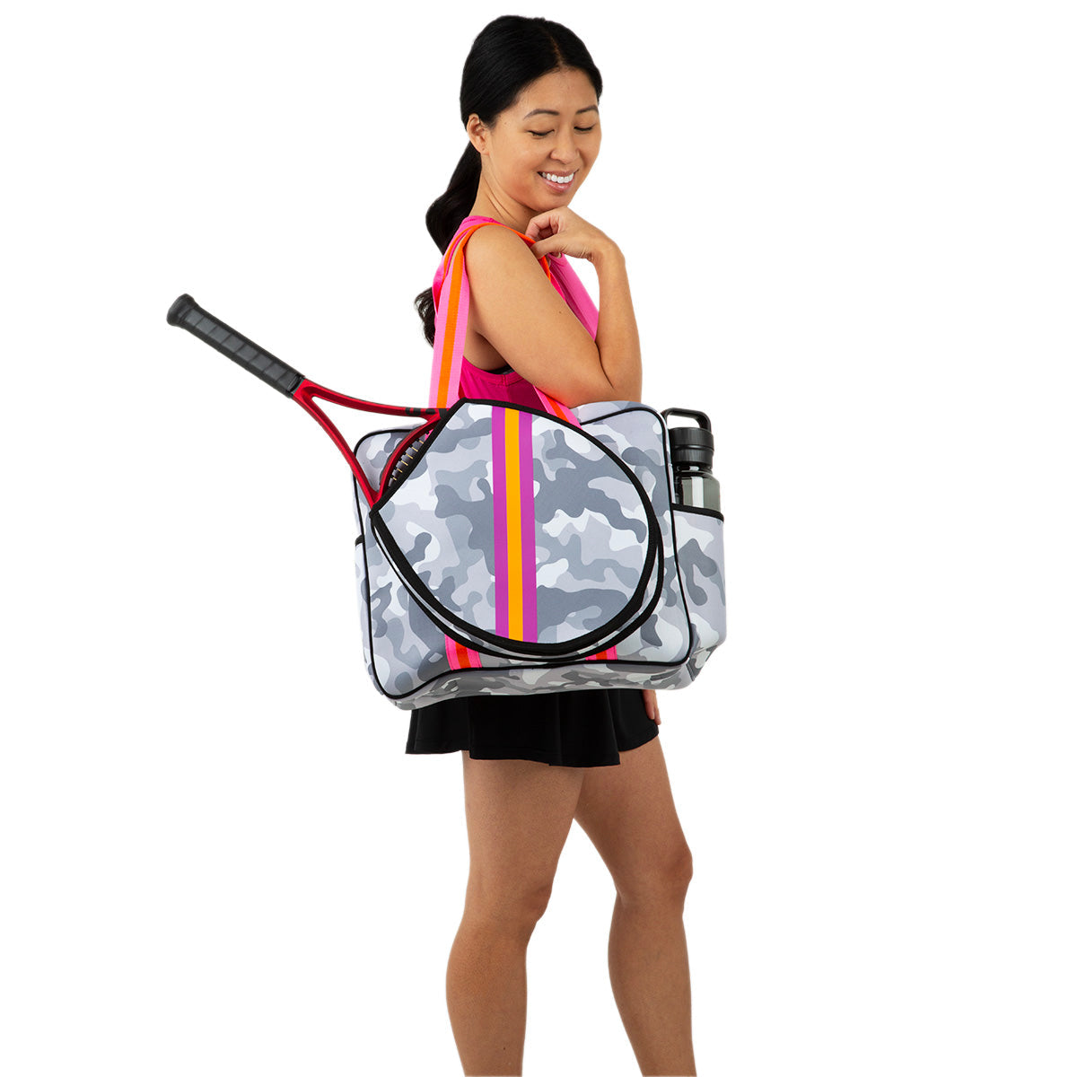 A woman in athletic wear holds the Queen of the Court Hampton Tennis Bag—a gray camo neoprene tennis bag with pink and yellow stripes—carrying a racket and two water bottles, smiling over her shoulder against a white background.