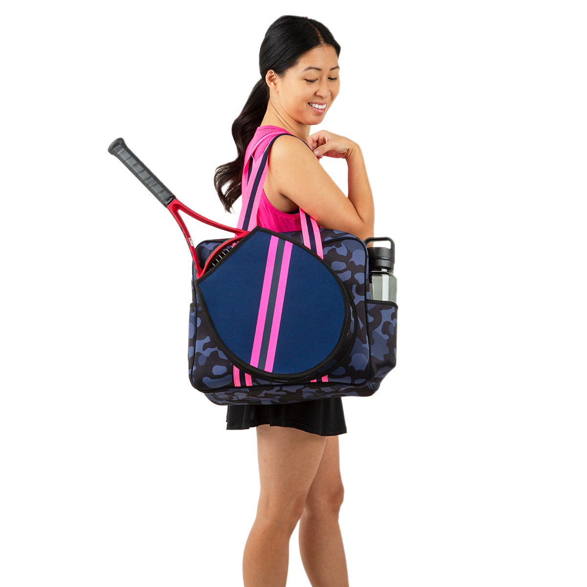 A woman in athletic wear smiles, holding a Queen of the Court Hampton Tennis Bag—navy blue and pink—over her shoulder. The neoprene bag holds a racket and silver water bottle as she stands against a plain white background.
