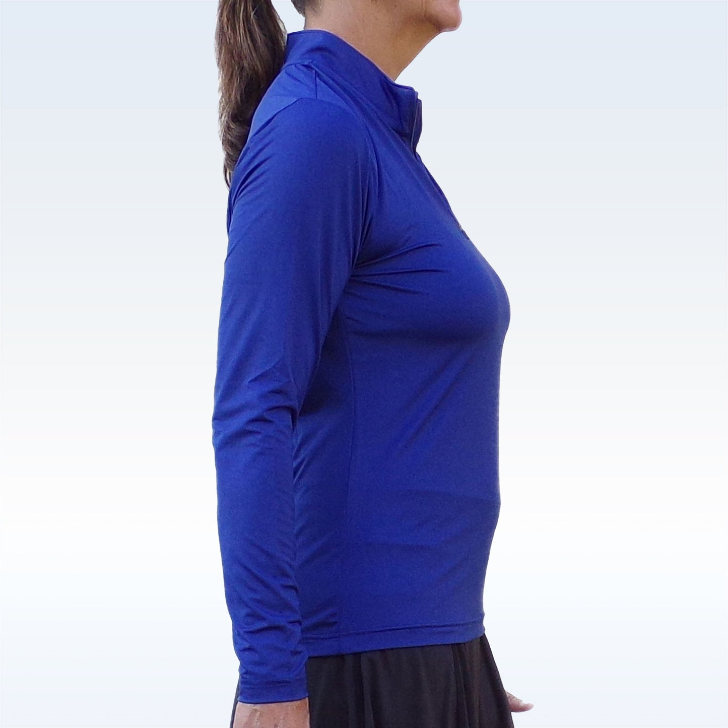 A woman with a ponytail and skirt stands sideways against a light gradient background, wearing the Pickleball Bella PB 1/4 Zip Long Sleeve Navy top featuring a high collar and SPF 50+ UV protection. Her face is not fully visible.
