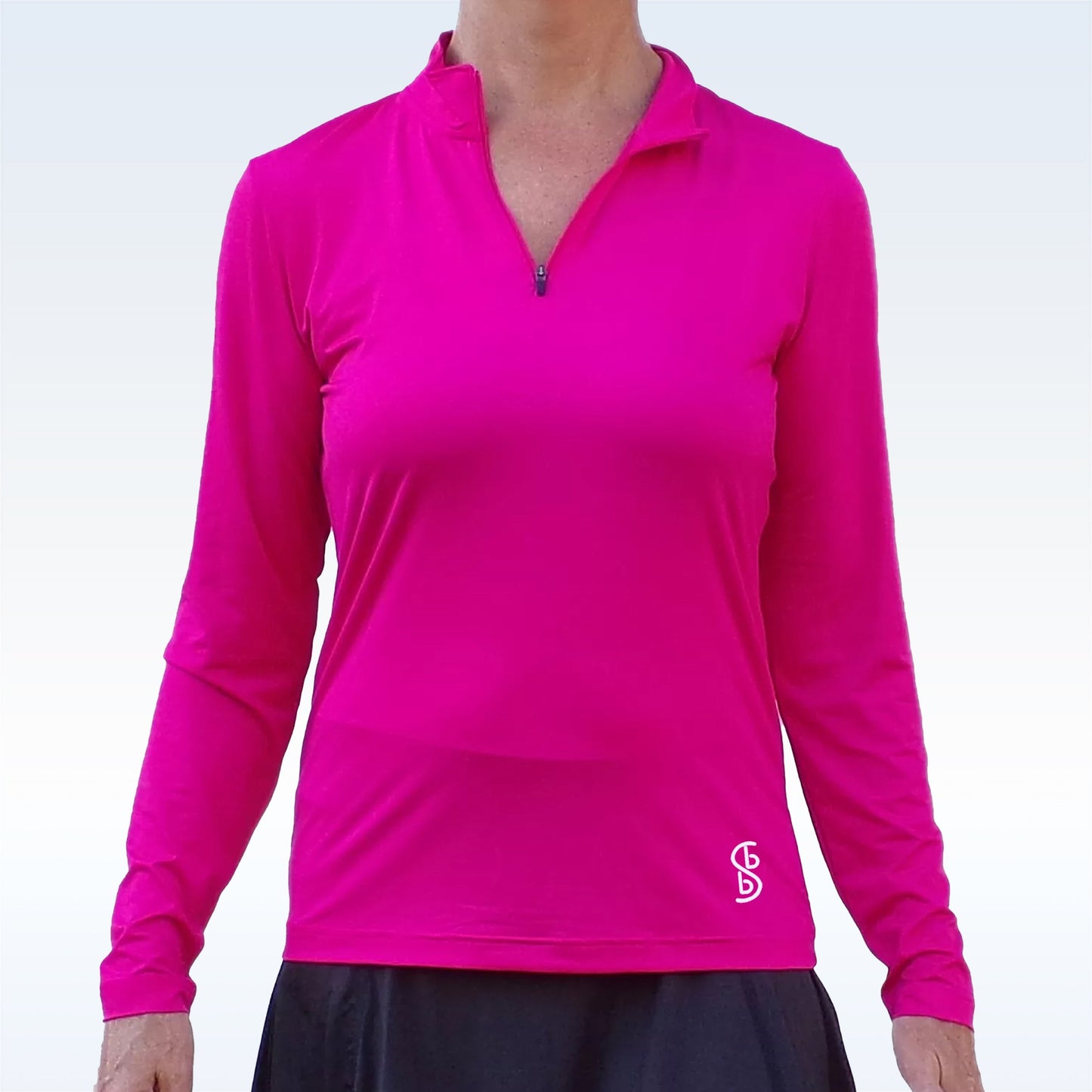 A woman models the Pickleball Bella PB 1/4 Zip Long Sleeve Pink—a bright pink, moisture-wicking top with a white logo on the lower left—paired with a black skirt. The image is cropped at her neck and above the knees against a plain light background.