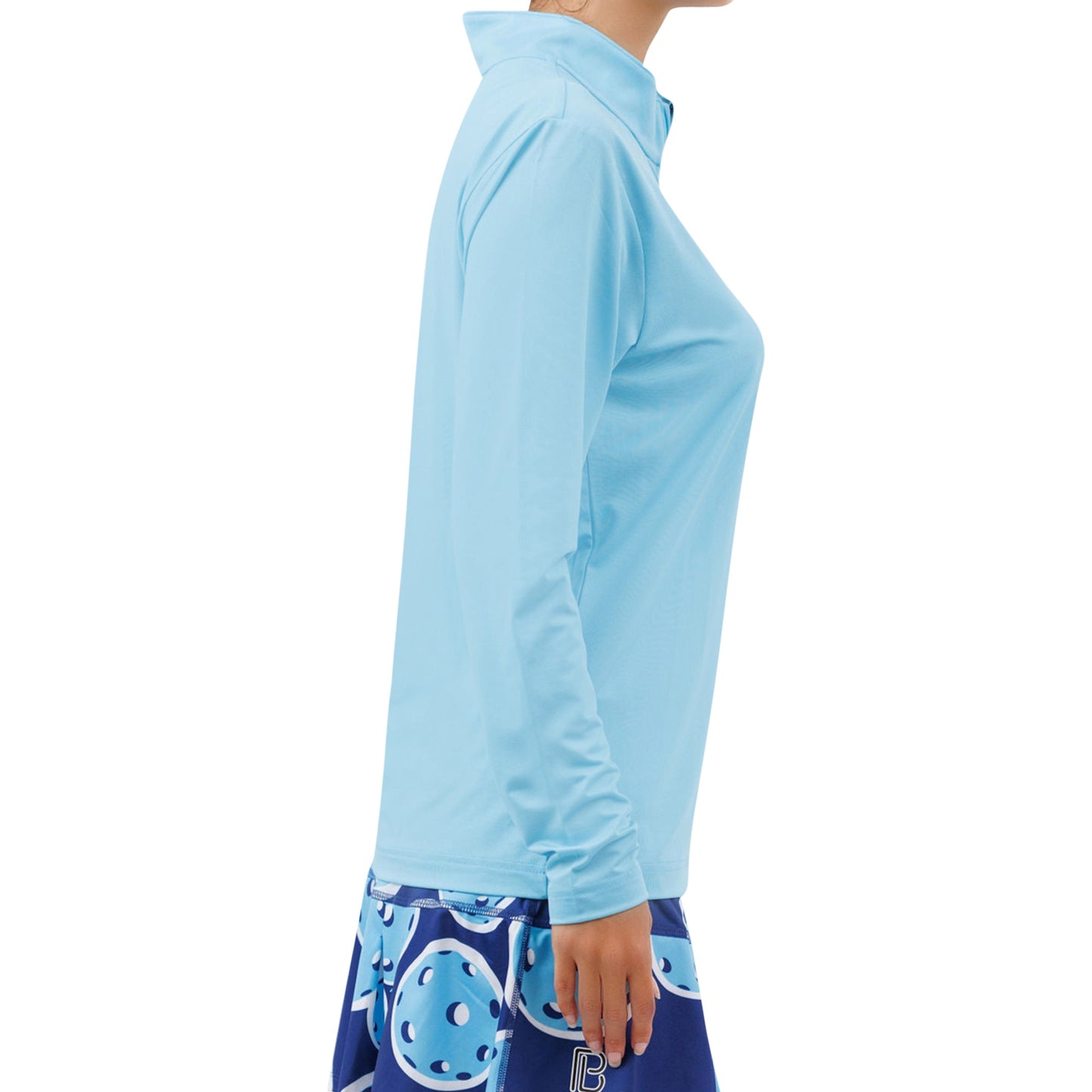 A person is shown in profile wearing the Pickleball Bella PB 1/4 Zip Long Sleeve Sky Blue, made from breathable fabric. They pair it with a blue skirt featuring abstract blue and white patterns. The image is cropped at the neck and mid-thigh.