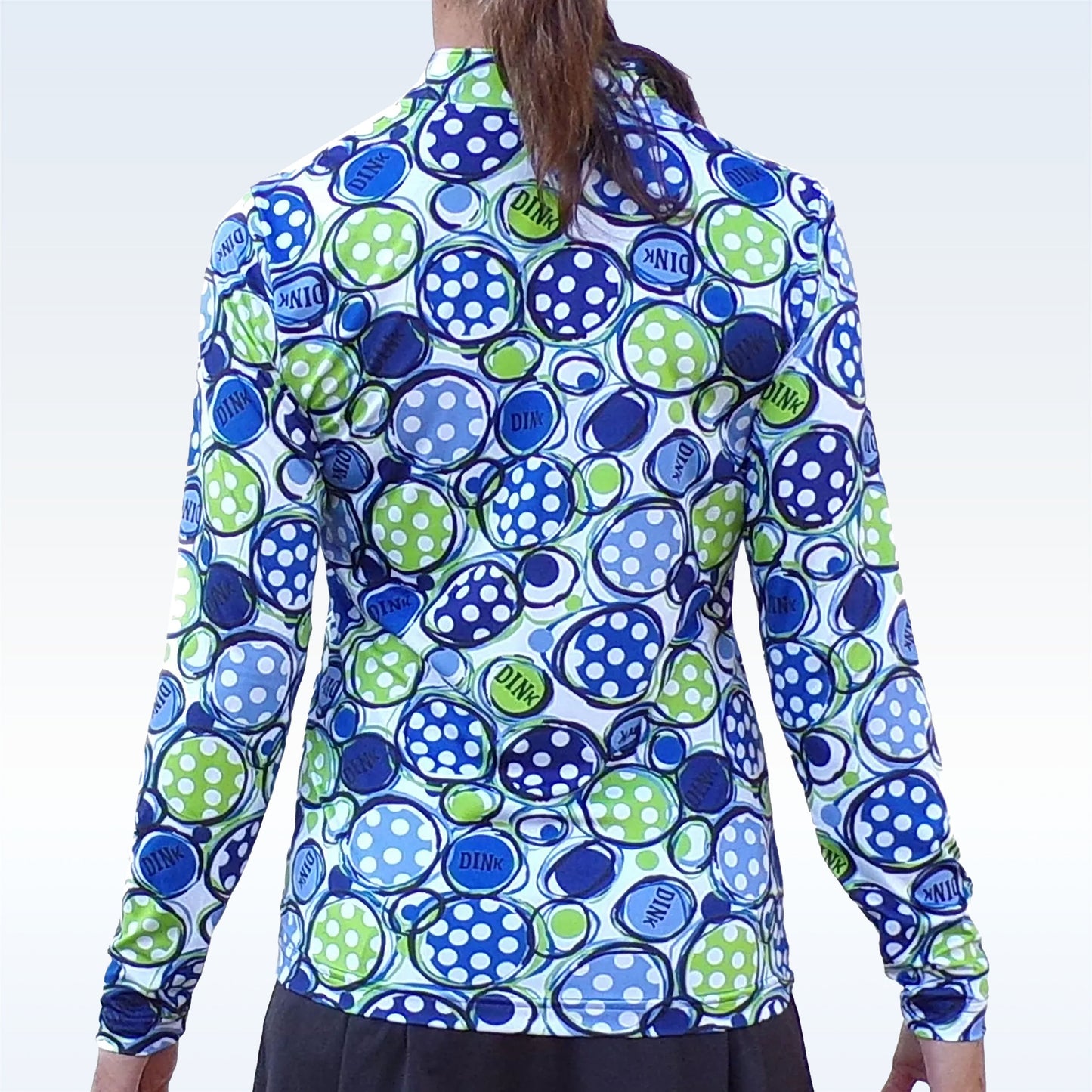 A person with long brown hair wears the Pickleball Bella PB 1/4 Zip Long Sleeve Dink 1, featuring a vibrant blue, green, and white pickleball design, SPF 50+ UV protection, and playful style shown from the back.