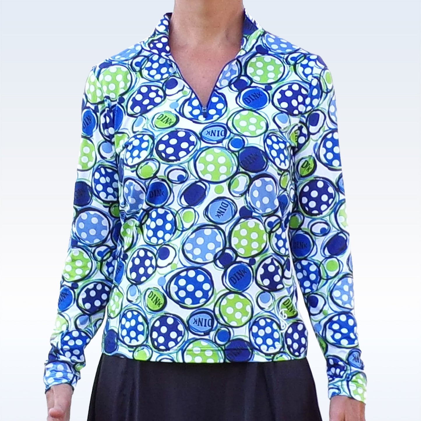 A person wears the Pickleball Bella PB 1/4 Zip Long Sleeve Dink 1 shirt, featuring blue, green, and white pickleball designs and a V-neckline. Paired with a black skirt, it delivers moisture wicking comfort and SPF 50+ UV protection.