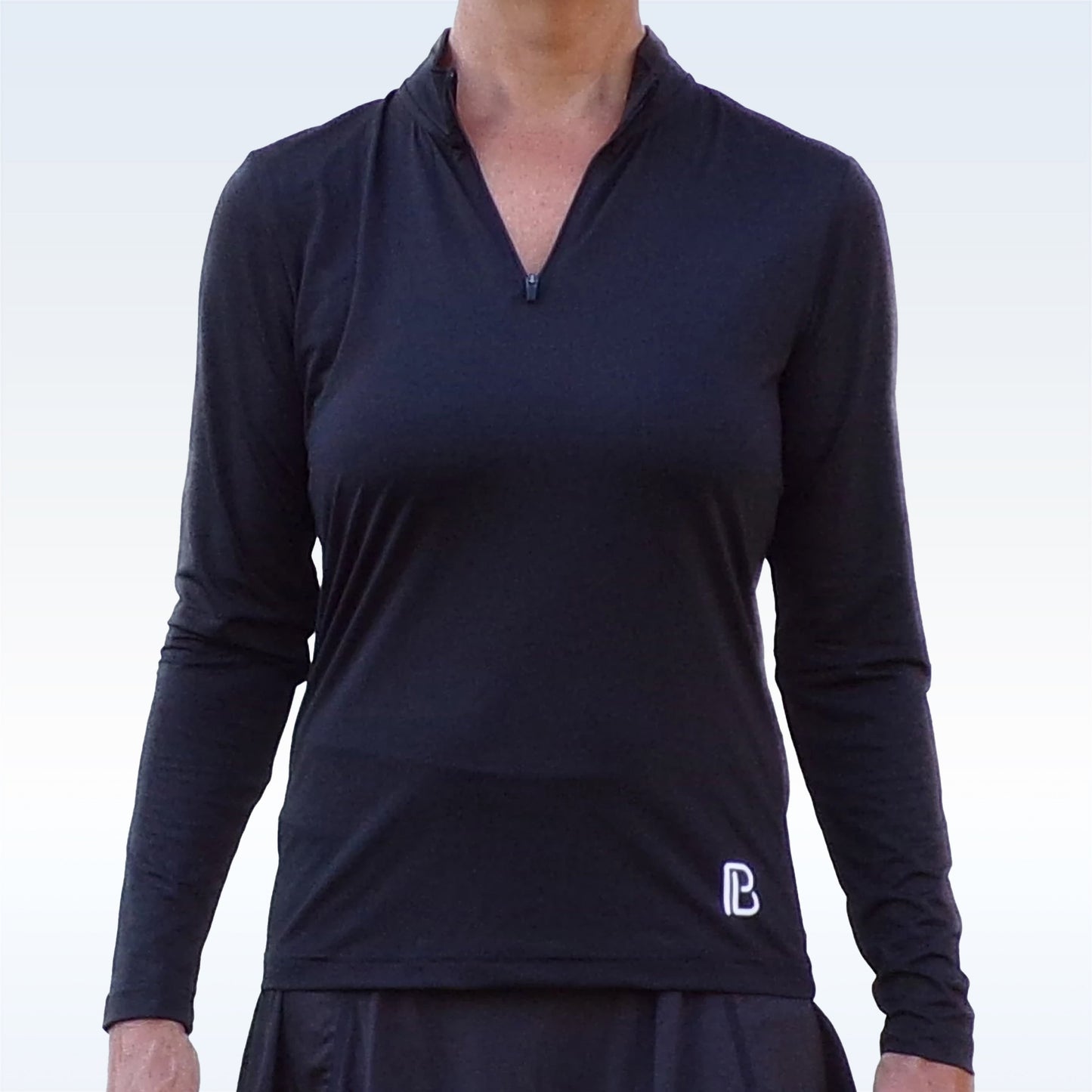 A person wears the Pickleball Bella PB 1/4 Zip Long Sleeve Black top, featuring moisture-wicking fabric and a small white logo on the lower left, against a plain light background. The head is not visible.