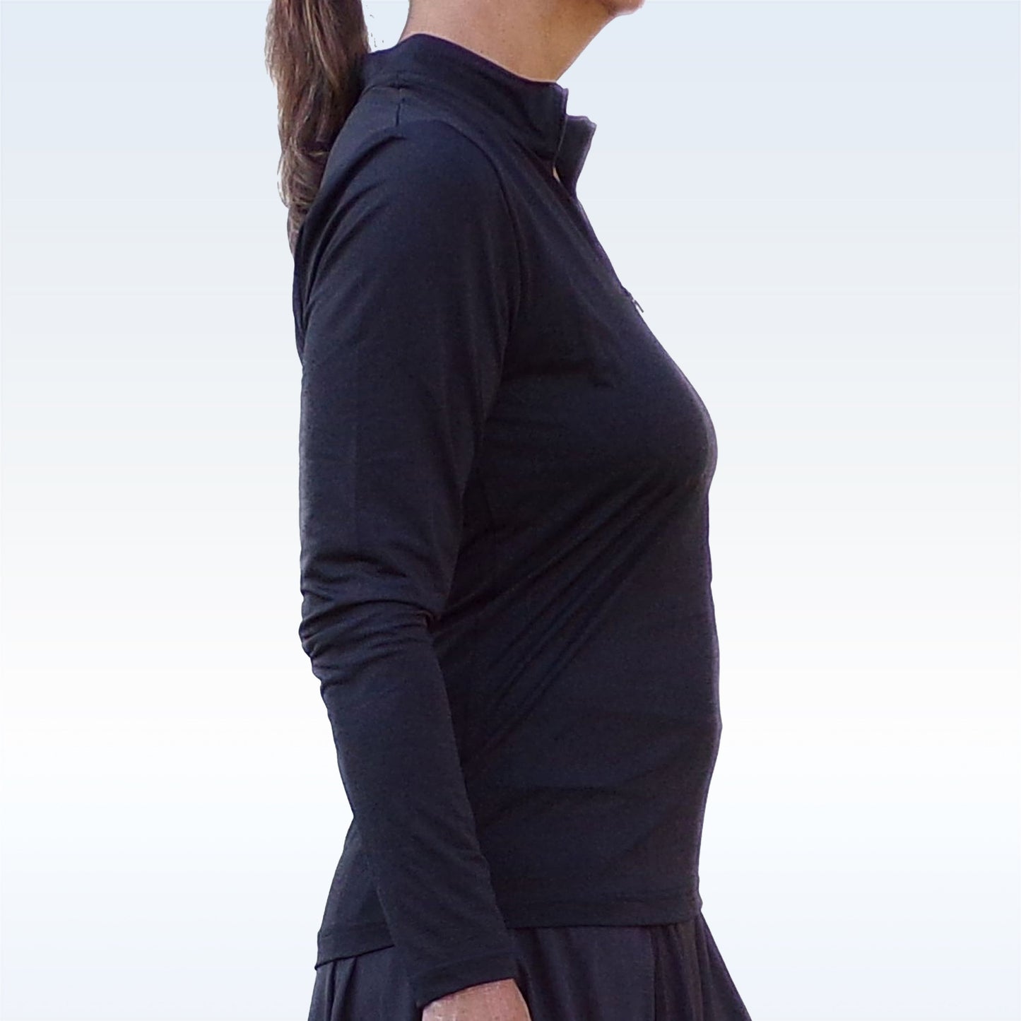 A person stands in profile facing left, wearing the Pickleball Bella PB 1/4 Zip Long Sleeve Black top paired with a black skirt. Their brown hair is tied back in a ponytail against a simple, light gradient background.