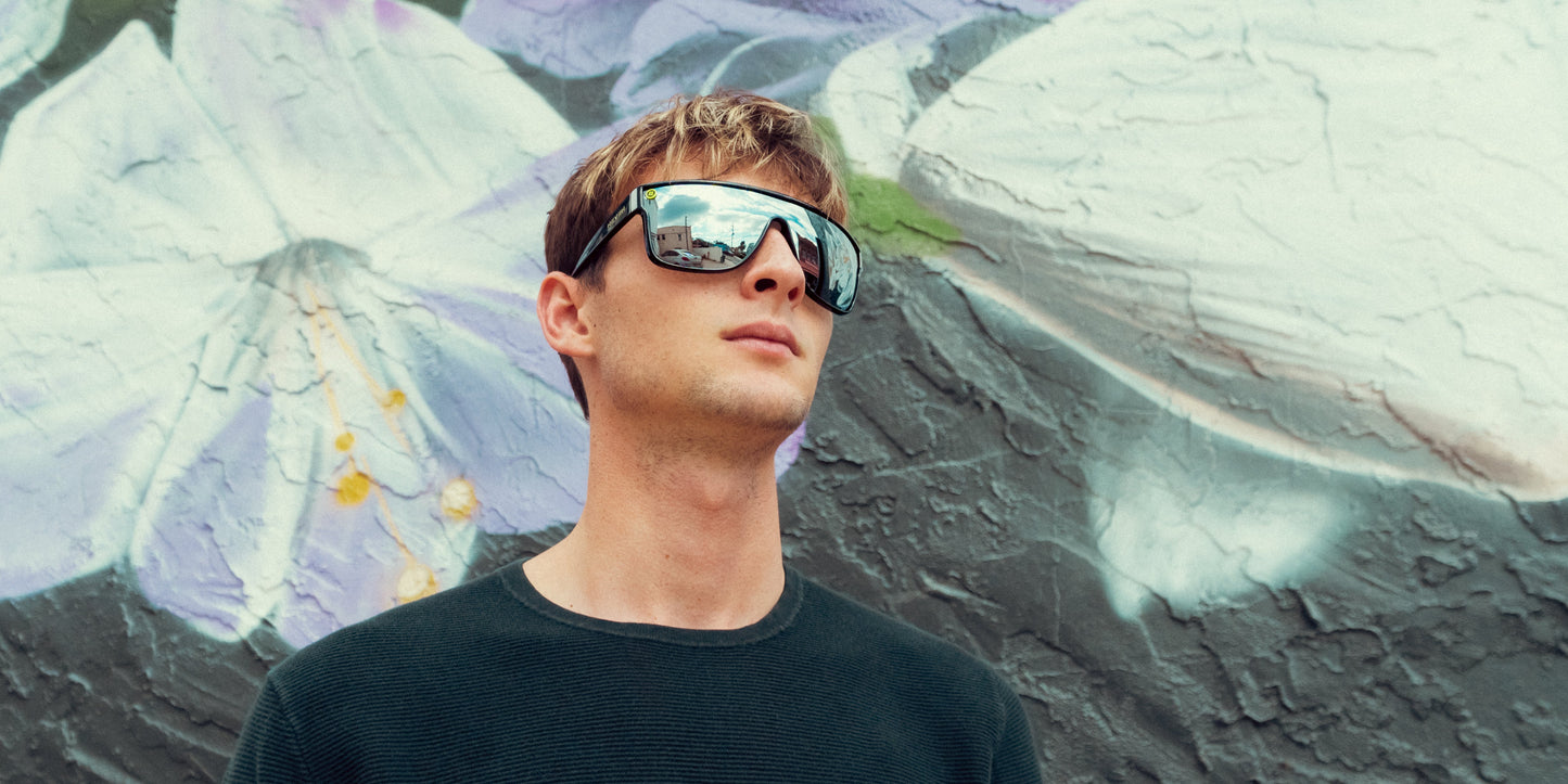 A young man wears Neven Eyewear 924 polarized sunglasses with stainless steel frames and UV400 lenses, reflecting city streets, as he stands before a textured mural of large white and purple flowers.