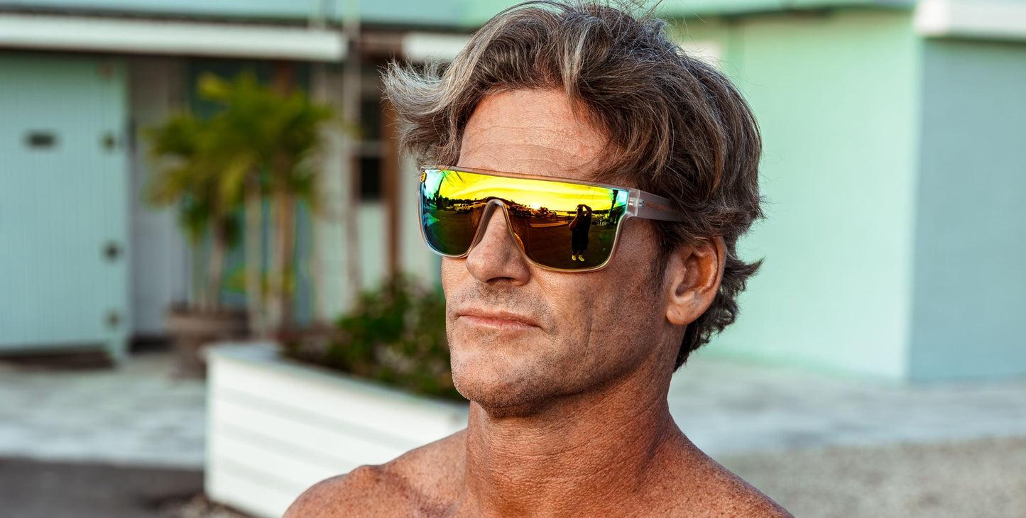 A middle-aged man with wavy brown hair and tanned skin wears Neven Eyewear’s Byron sunglasses with polarized UV400 protection. He stands shirtless outdoors, a modern light blue building and tropical plants blurred behind him.