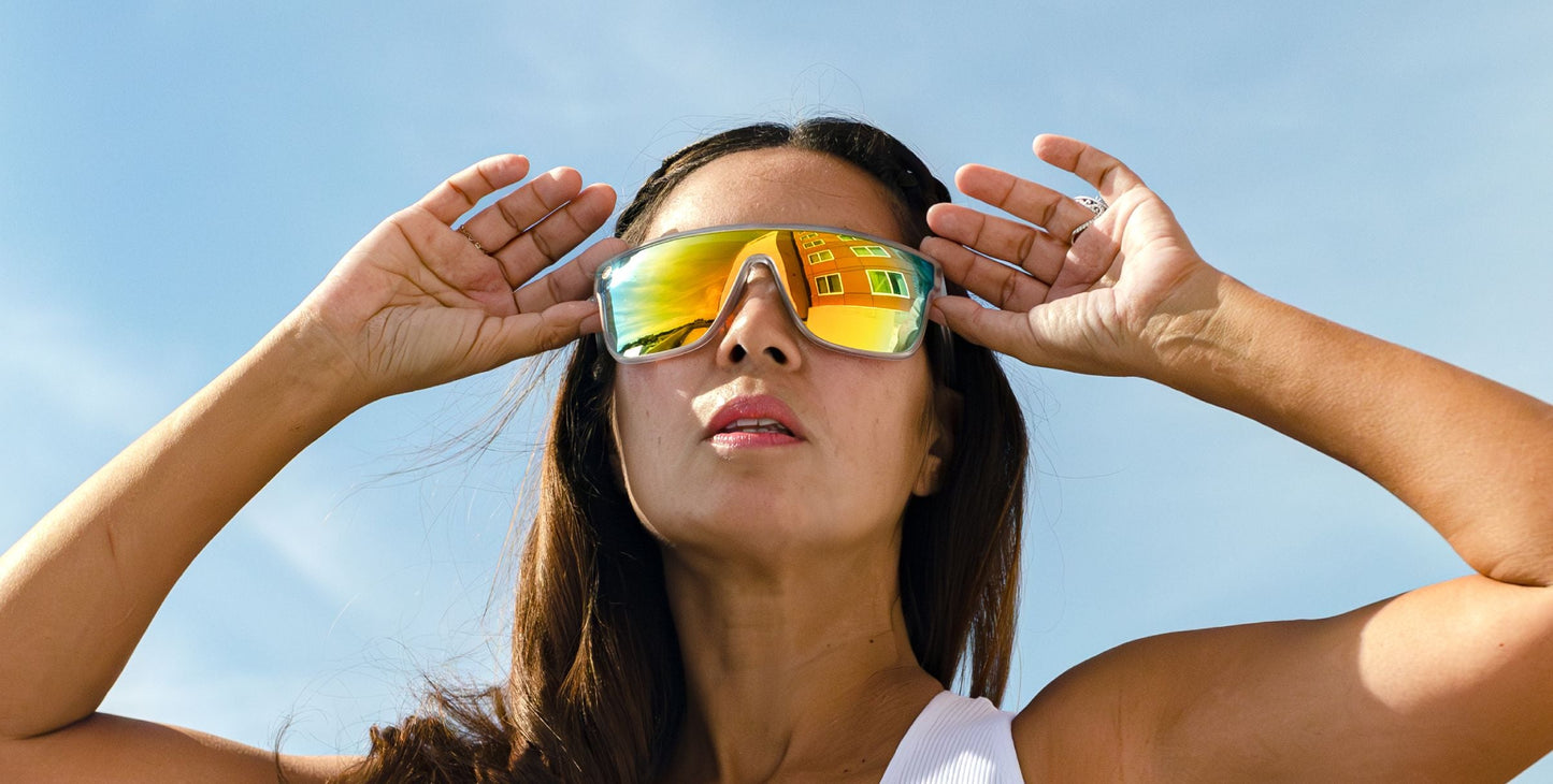 A person with long brown hair adjusts their Neven Eyewear Byron sunglasses with polarized UV400 protection. The large, reflective lenses mirror a bright yellow building as they stand outside in a white sleeveless top beneath a clear blue sky.