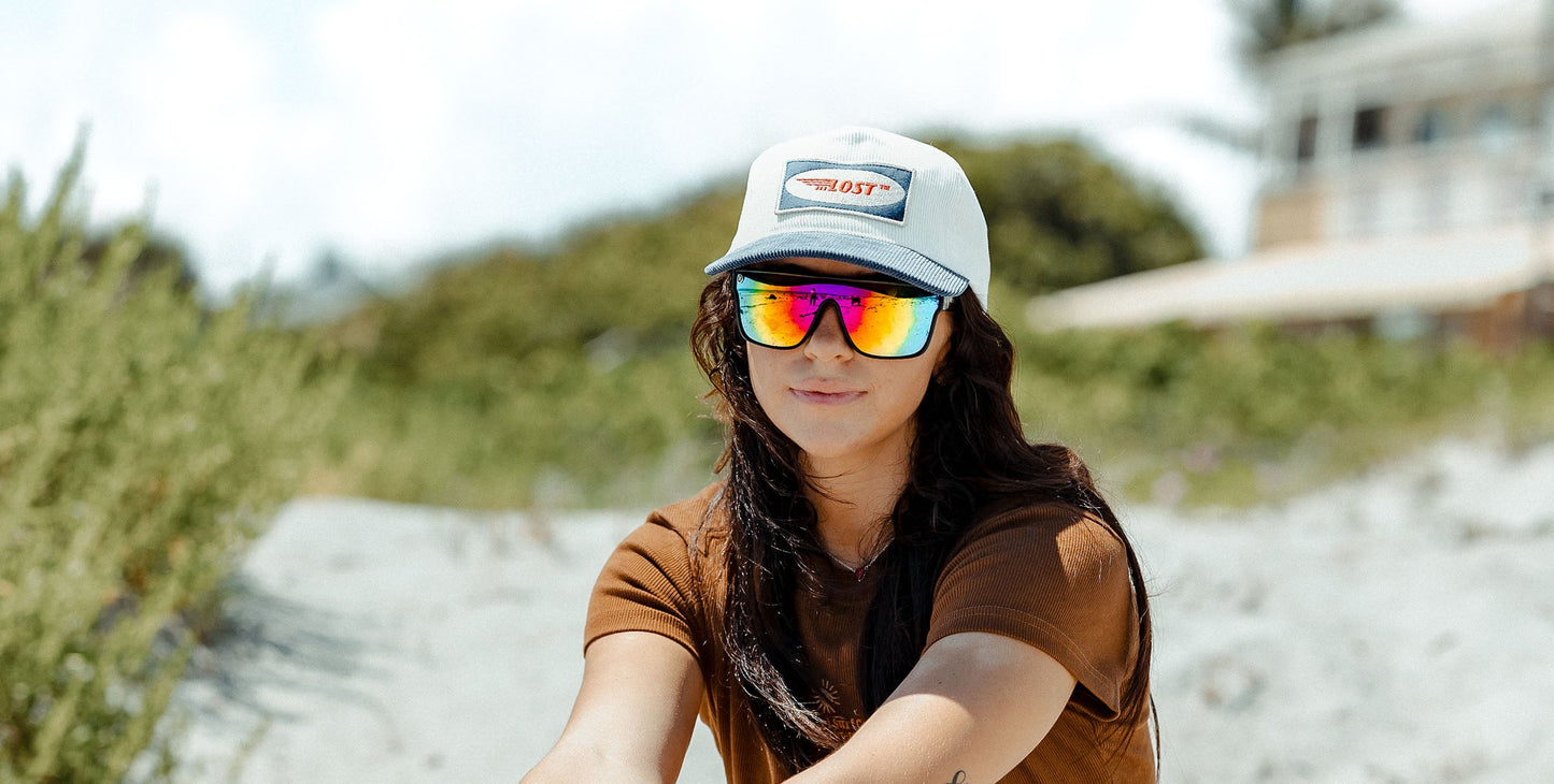 A woman with long dark hair wears the Noosa reflective polarized sunglasses by Neven Eyewear, a brown shirt, and a light gray cap with a LOST patch while outdoors in a sunny setting with greenery and a blurred building behind her.