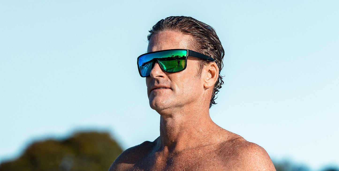 A middle-aged man with slicked-back brown hair and tanned skin stands shirtless outdoors, wearing Neven Eyewear’s Valley polarized sunglasses (UV400). Blue sky and green trees are blurred in the background.