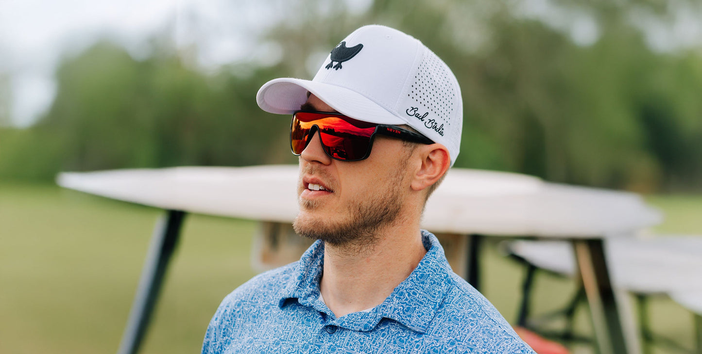 A man with a short beard wears the "Well Done" red polarized sunglasses by Neven Eyewear, featuring UV400 protection. He pairs them with a white cap, blue patterned shirt, and stands outdoors among trees and picnic tables.