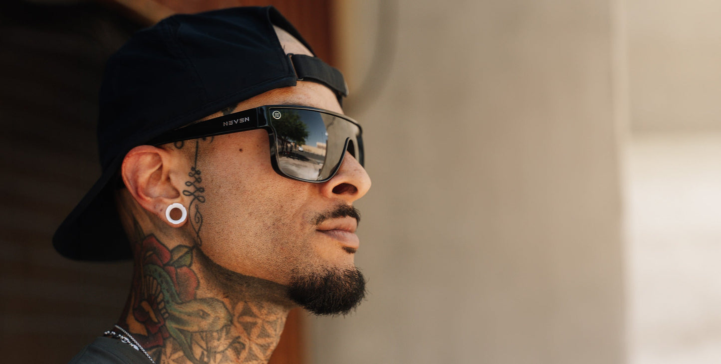 A man with a goatee, stretched earlobes, colorful neck tattoos, and facial piercings looks to the side. He sports a black backward cap and wears Neven Eyewear's Wire sunglasses with polarized UV400 protection. The background is blurred.