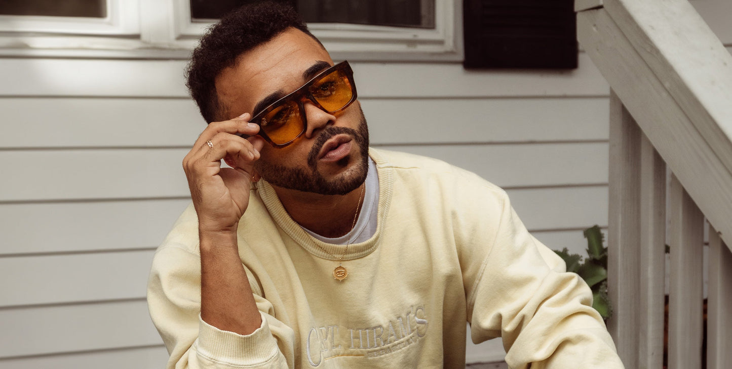 A man with short curly hair, beard, and mustache sits on outdoor steps in front of a white house, wearing a pale yellow sweatshirt. He holds Neven Eyewear's Amar tortoise shell sunglasses stylishly in one hand.