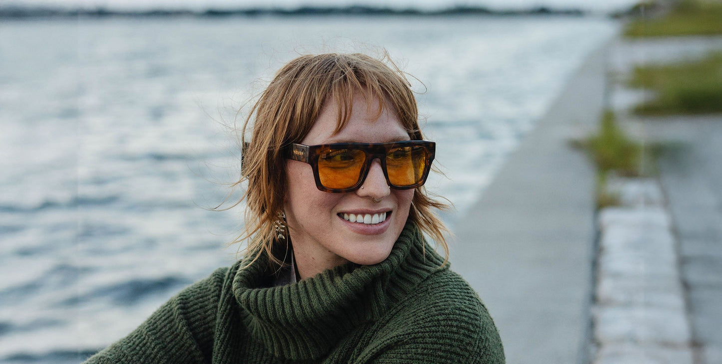 A person with short, tousled light brown hair smiles while wearing Neven Eyewear's Amar fashion frames with amber lenses, a green knit turtleneck, and dangling earrings beside a waterfront and concrete walkway.