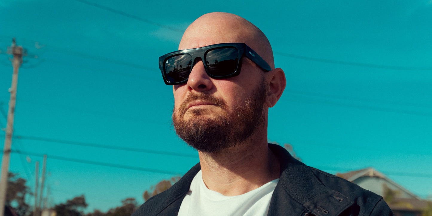 A bald man with a trimmed beard and mustache wears Neven Eyewear's Royce polarized sunglasses with UV400 protection and a dark jacket over a white shirt, standing outdoors under a bright blue sky with power lines and houses behind him.