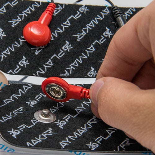 A hand attaches a red connector to a round silver snap on a black rectangular pad labeled iReliev PLAYMAKAR, part of the SPORT Muscle Stimulator with Explosive Strength Mode. The pad shows white logos; another connected pad is visible above.