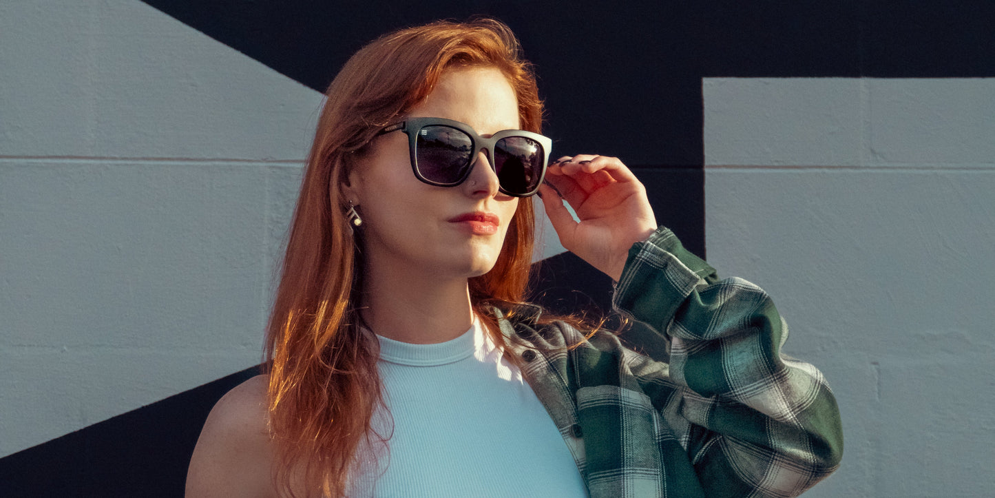 A woman with long red hair wears Neven Eyewear’s Lido sunglasses with matte black frames and a white sleeveless top, her green plaid shirt over one shoulder. She stands by a bold geometric wall, adjusting the polarized UV400 shades.
