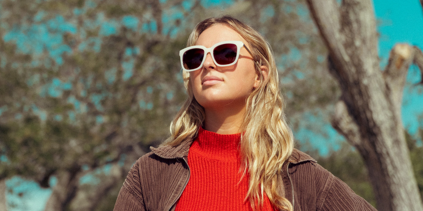 A woman with long blonde hair wears Neven Eyewear’s Sand Dollar sunglasses—oversized white frames with gold metal arms—paired with a red sweater and brown corduroy jacket, standing confidently outdoors under a blue sky.