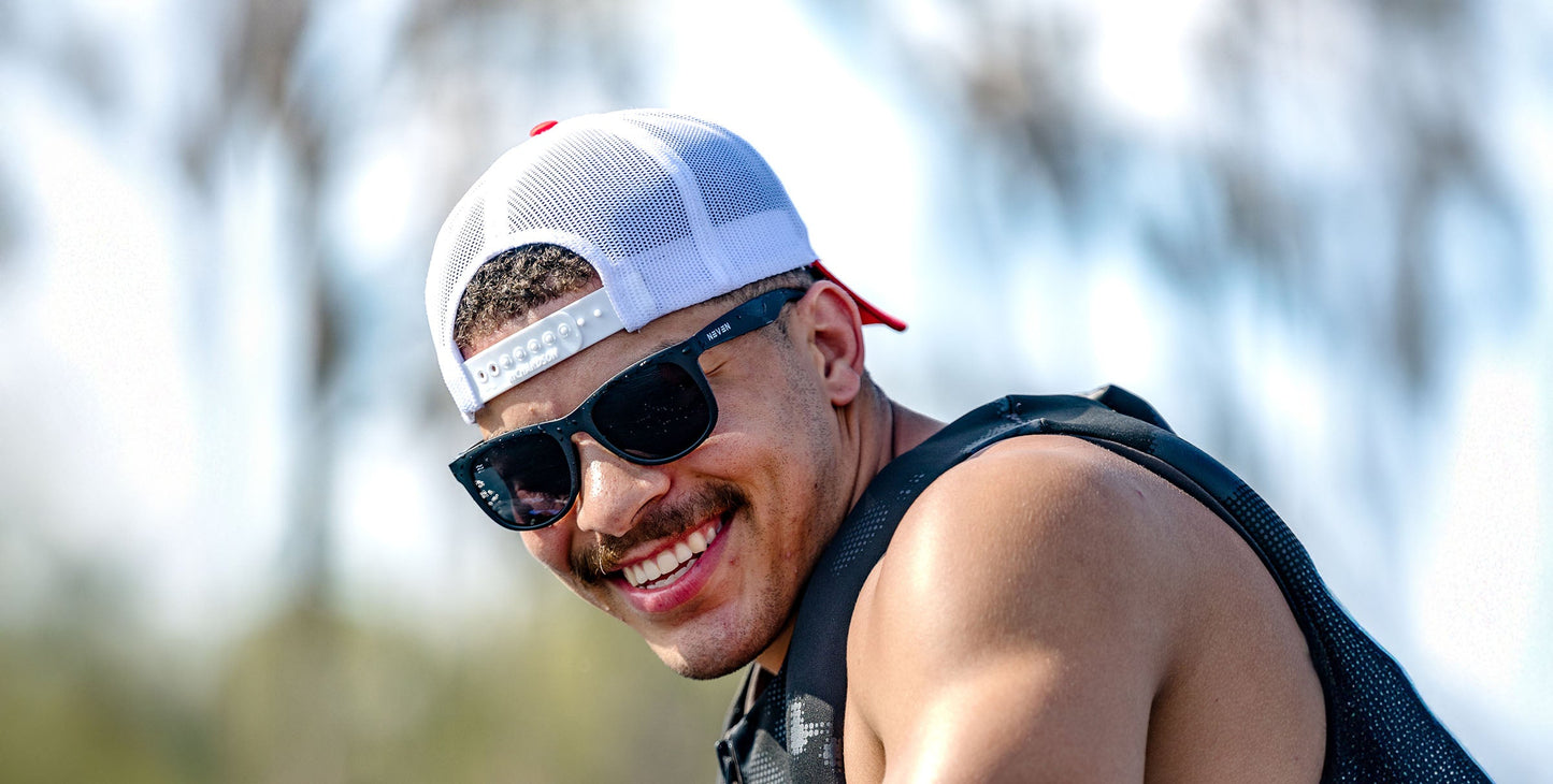 A young man with a mustache and goatee smiles at the camera, wearing Neven Eyewear's Classic black polarized sunglasses with UV400 protection, a white cap with a red brim, and a sleeveless black shirt against a softly blurred outdoor backdrop.