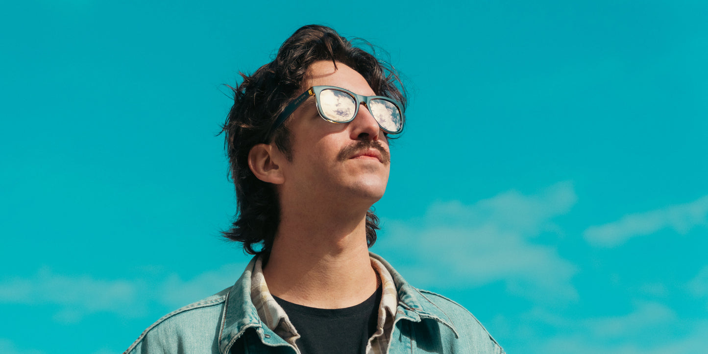 A person with wavy dark hair and a mustache gazes upward against a bright blue sky, wearing Neven Eyewear's Sundown polarized sunglasses, which reflect clouds. Soft clouds are visible in the background. They wear a light denim jacket and dark shirt.