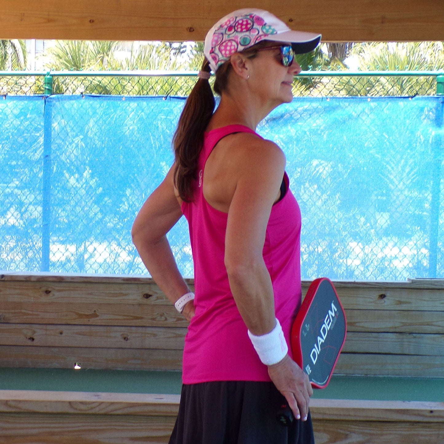A woman wearing the Pickleball Bella PB Racerback Tank Pink, a patterned cap, sunglasses, and wristbands stands on a pickleball court with a DIAD3M paddle. She faces right with her left hand on her hip; a wooden bench and blue fence are behind her.