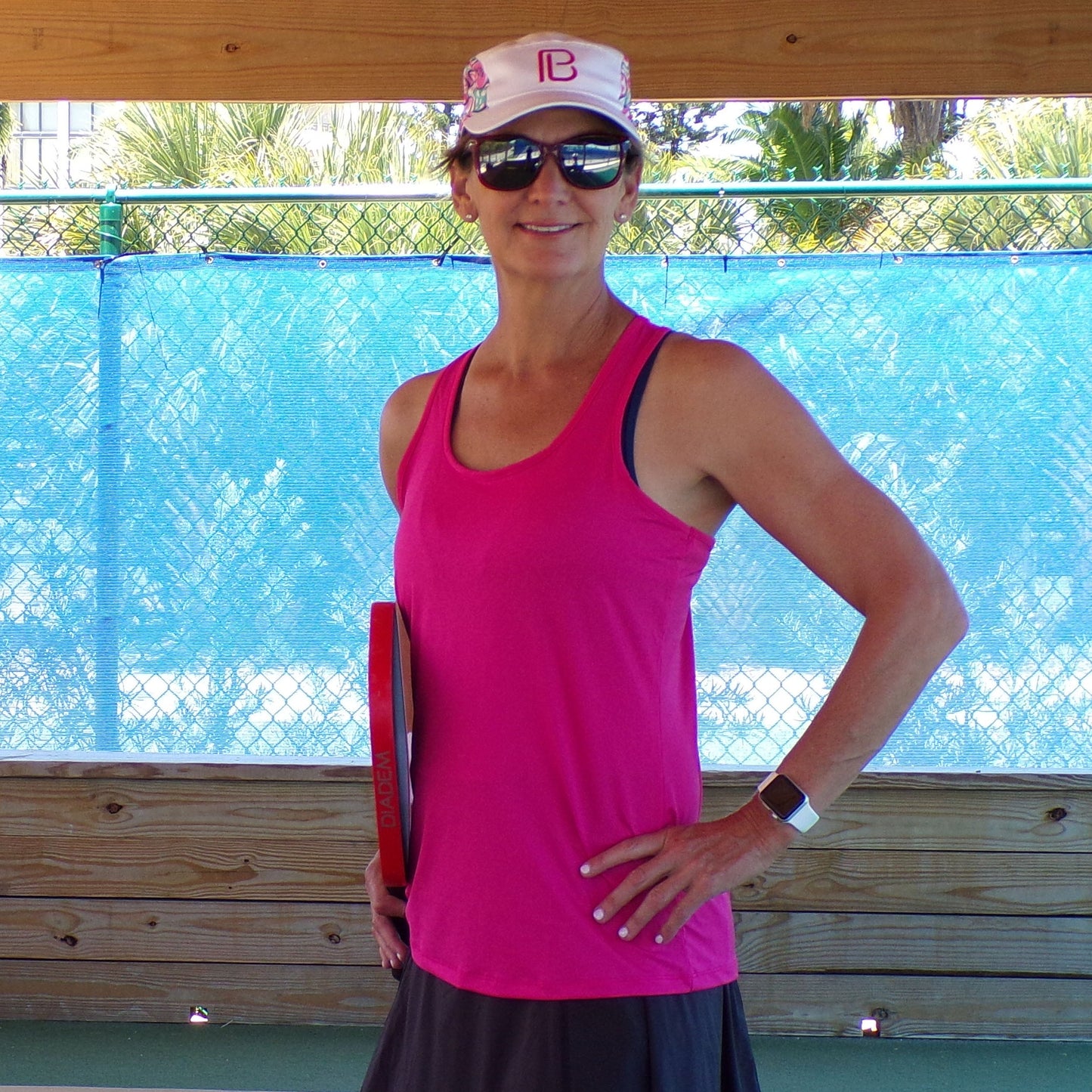 A smiling woman in a Pickleball Bella PB Racerback Tank Pink with SPF 50+ UV protection, black skirt, white cap, and sunglasses stands holding a red pickleball paddle by a blue fence and wood barrier, palm trees in the background.